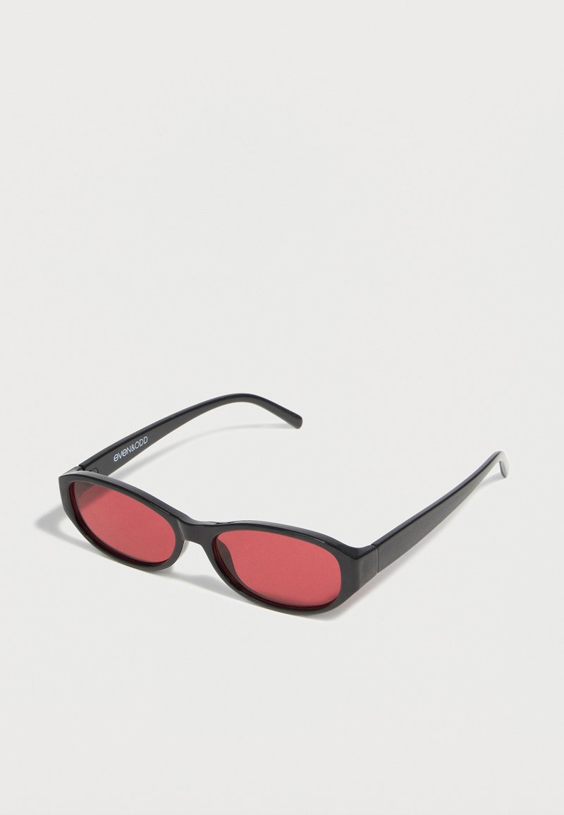 Μαύρα οβάλ γυαλιά ηλίου με λεπτούς βραχίονες και κόκκινα φακούς, τοποθετημένα πάνω σε λευκή επιφάνεια.