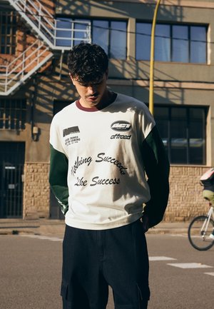 Young man with curly hair wearing a white and green long-sleeve shirt with text, standing on a street near a building and a cyclist passing by.