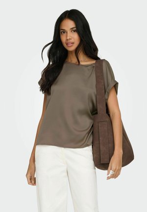 Kvinde med langt mørkt hår iført en løstsiddende brun kortærmet top, hvide bukser og bærende en stor brun skuldertaske.