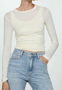 Top ajustado de manga larga en amarillo claro, superpuesto a una camiseta blanca transparente. Combinado con jeans de tiro alto en azul, que presentan una textura de mezclilla clásica.