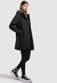 Veste noire imperméable avec capuche, fermeture éclair à l'avant et poches latérales, associée à des leggings noirs et à des baskets montantes noires avec des semelles blanches.