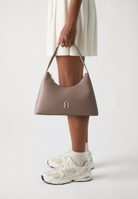 Borsa a spalla in pelle beige con un design curvo, logo in hardware dorato e una sola tracolla. La borsa è tenuta da una persona che indossa un'abito bianco e sneaker.