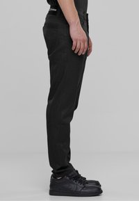Čierne slim-fit džiny s hladkou textúrou, so bočnými vreckami a logom. V páre s čiernymi teniskami. Bočný profil.