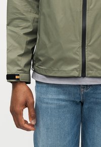 Άνδρας που φοράει ελιάς πράσινο σακάκι με μαύρο φερμουάρ και ρυθμιζόμενο μανίκι, γκρι πουκάμισο από κάτω και μπλε τζιν, στέκεται με το αριστερό χέρι χαλαρό.