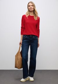 Roter Pullover mit langen Ärmeln, kombiniert mit dunkelblauen Schlagjeans. Das Outfit umfasst eine beigefarbene Wildledertasche und weiße Sneakers mit strukturierten Details.
