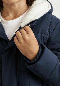 Mornarska puhovka z mehkimi belimi flis podloženimi kapuco, kvadratnim dizajnom in rebrastimi manšetami, zaprta z zadrgo, s roko, ki drži ovratnik.