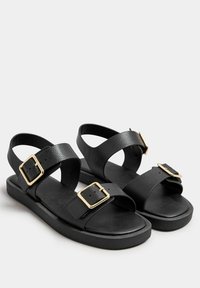 Mustat nahkasaappaat, joissa on kaksi leveää hihnaa kultaisilla soljilla, tasainen pohja ja teksturoitu pinta lisäpidon ja mukavuuden takaamiseksi.