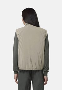 Gilet imbottito in beige chiaro, con colletto alto, senza maniche e orlo elastico. Indossato sopra una maglietta a maniche lunghe verde scuro.