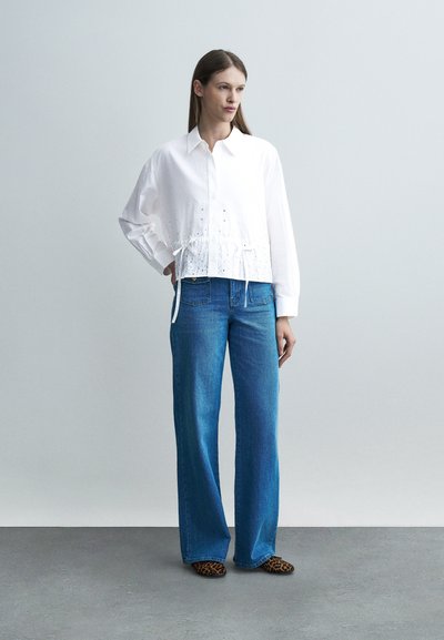 Femme debout, main sur la hanche, portant une chemise blanche à manches longues, un jean bleu taille haute à jambes larges et des chaussures imprimé léopard sur un sol gris.