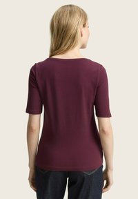 Frau mit blonden Haaren, die ein schlichtes dunkelviolettes T-Shirt mit kurzen Ärmeln und dunkelblaue Jeans trägt, von hinten vor einem hellen Hintergrund gezeigt.
