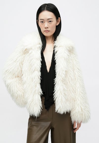 Béžový načechraný kabát s dlhými, štruktúrovanými chlpmi, širokým golierom a spustenými ramenami, skombinovaný s čiernym plisovaným topom a hnedými koženými nohavicami.