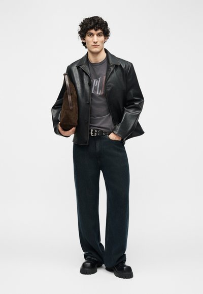 Νεαρός με σγουρά μαλλιά που φοράει μαύρο δερμάτινο μπουφάν, γραφικό μπλουζάκι, φαρδιά μαύρα τζιν, χοντρούς μαύρους παπούτσια, κρατώντας καφέ σακίδιο από σουέντ.