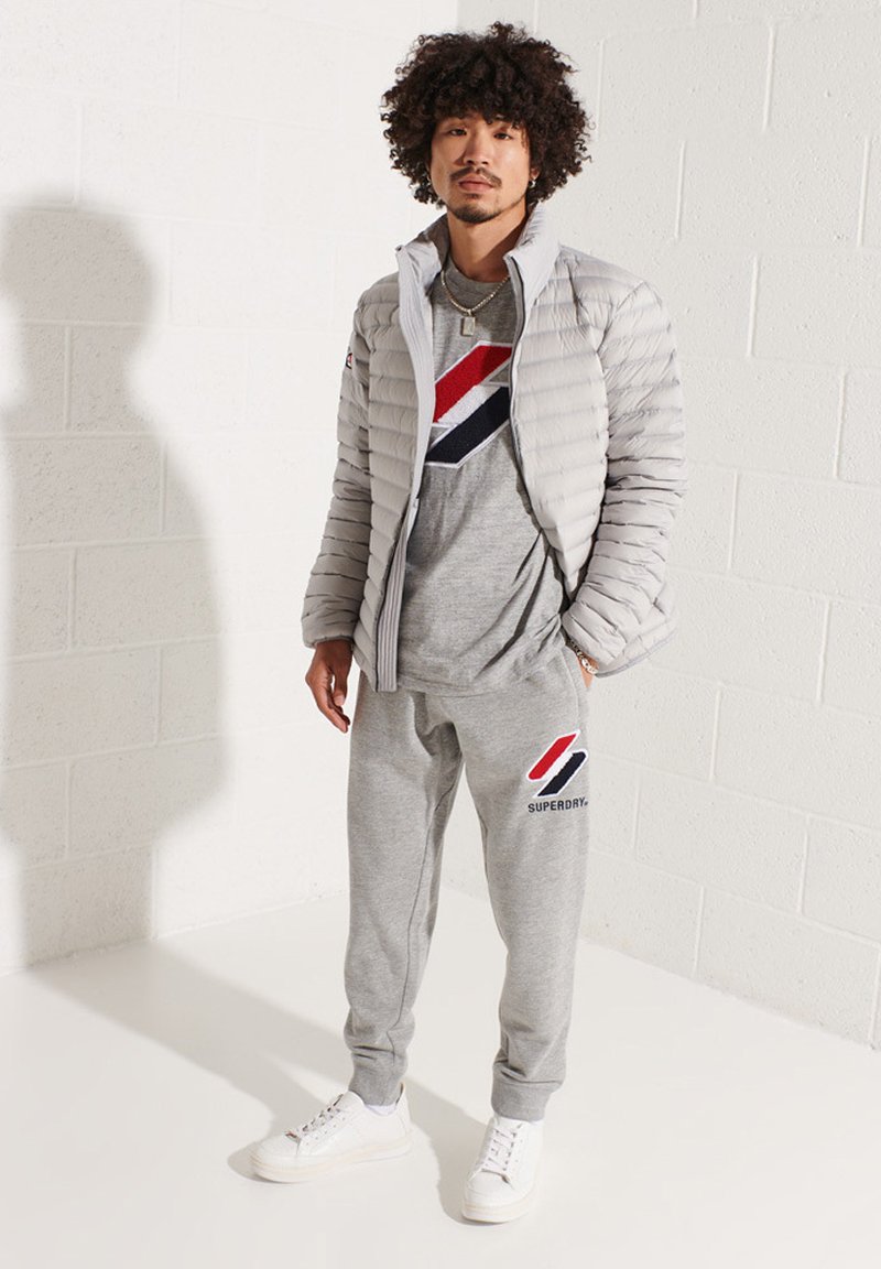 Doudoune grise avec un design matelassé horizontal, pantalon de survêtement gris assorti présentant un logo en blocs de couleur rouge et bleu, et des sneakers blanches.