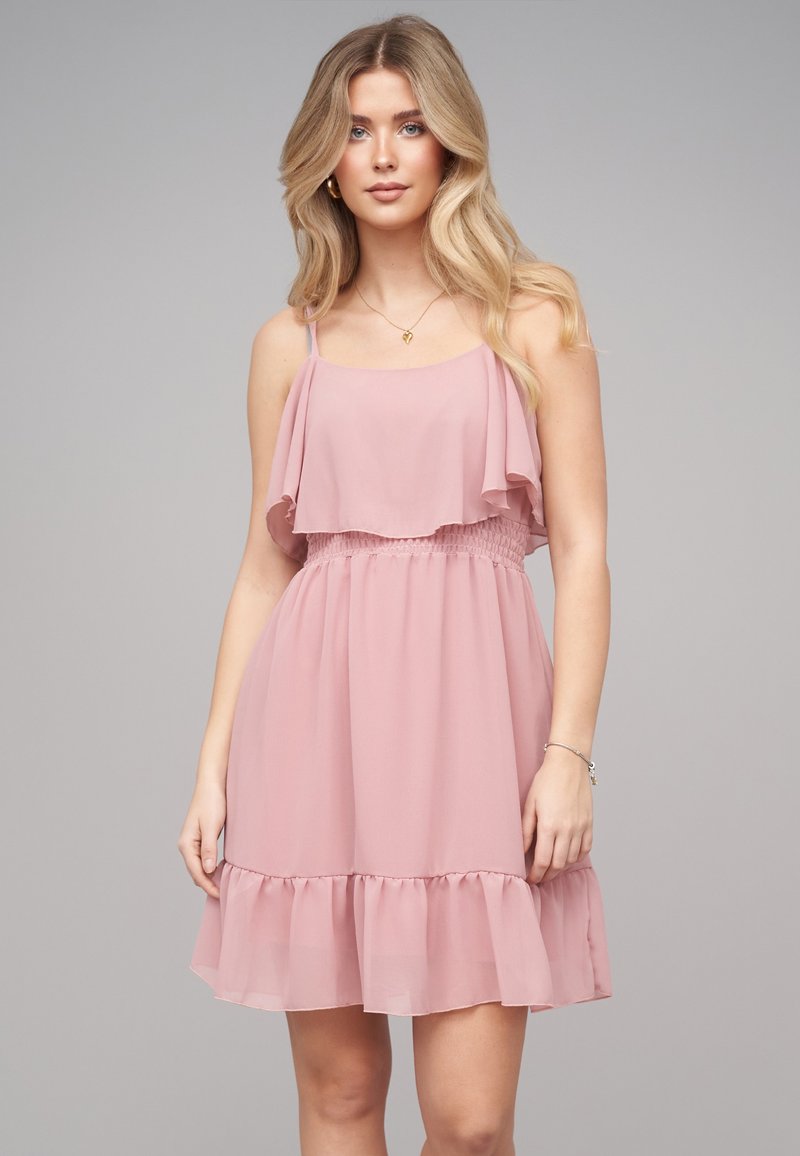 Jeune femme aux longs cheveux blonds portant une robe rose clair sans manches à volants sur un fond gris uni.