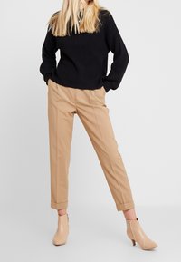 Black textured sweater with wide sleeves, paired with beige tailored trousers and light beige ankle boots with a small heel.