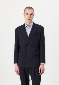 Marineblauwe dubbele blazer in een gladde stof, met twee zwarte knopen, notch revers en geflapte zakken. Gelaagd over een lichtblauw shirt.