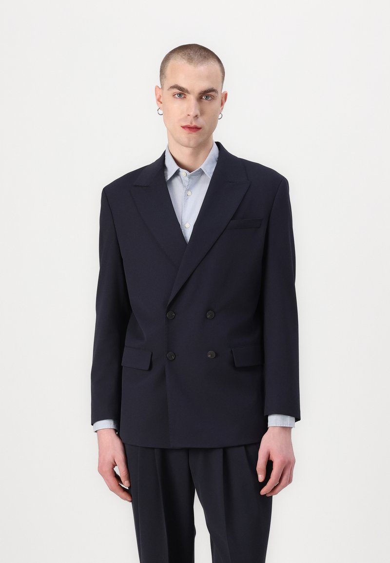 Marineblauwe dubbele blazer in een gladde stof, met twee zwarte knopen, notch revers en geflapte zakken. Gelaagd over een lichtblauw shirt.