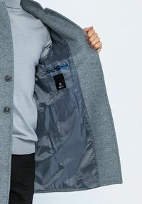 Cappotto di lana grigio con chiusura a bottoni e due tasche interne. Foderato in morbido tessuto grigio, una tasca presenta un accento a motivo blu.