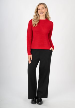 Roter Strickpullover mit gerippter Textur und langen Ärmeln, kombiniert mit schwarzen weiten Hosen und schwarzen Schuhen, vor einem schlichten Hintergrund stehend.