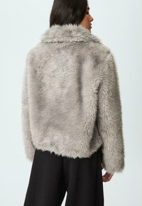 Donna di profilo che indossa una giacca in pelliccia sintetica grigia chiara e pantaloni neri, su uno sfondo chiaro e semplice.
