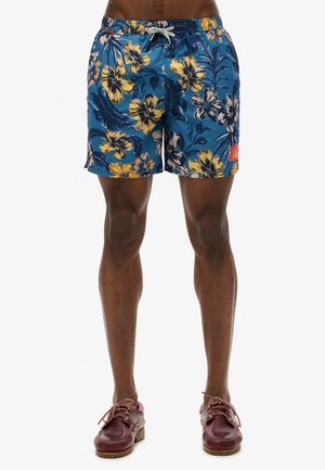 Shorts de bain floraux bleus avec des fleurs jaunes et blanches, une taille élastique avec un cordon de serrage, et un accent de logo rouge sur le côté.