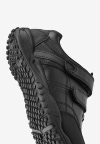 Chaussure de sport noire avec une semelle en caoutchouc texturé, une sangle en velcro, un matériau supérieur lisse et des coutures renforcées. La vue en angle montre les détails sur le côté.