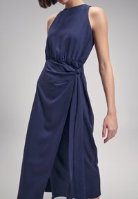 Navy blue sleeveless dress with a tied waist detail and draped fabric, featuring a long skirt and smooth texture.