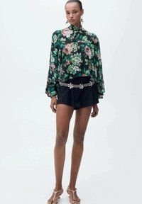 Blommig blus i svart med gröna och rosa blommor, med långa ärmar och hög hals, som är matchad med svarta shorts och ett dekorativt bälte.
