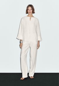 Haut en lin blanc avec un col rond et une fermeture à boutons, associé à un pantalon large assorti. Texture uniforme, design minimal et coupe décontractée.