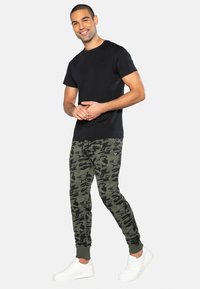 Schwarzes T-Shirt mit kurzen Ärmeln kombiniert mit grünen Camo-Jogginghosen. Die Jogginghosen haben Knöchelabschlüsse und ein kleines Logodetail. Weiße Sneakers runden den Look ab.