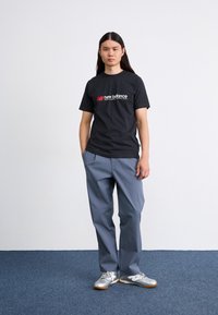 Sort bomulds t-shirt med et rødt og hvidt "New Balance" logo, parret med grå bukser og sølvfarvede træningssko på et blåt tæppe.