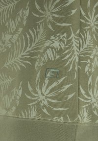 Tela verde con un patrón de hojas en tonos verdes claros, con un logotipo bordado de marca y detalles de costura visibles a lo largo de la parte inferior.