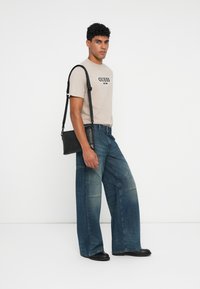 Hombre que lleva una camiseta beige de GUESS, jeans anchos desgastados, zapatos negros y lleva un bolso cruzado negro sobre su hombro.