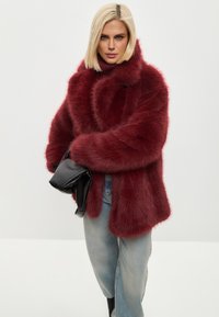 Abrigo de pelo sintético burdeos con un cuello alto, mangas largas y bolsillos laterales; combinado con jeans azul claro y un bolso negro.