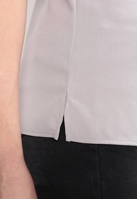 Blusa grigia chiara con una texture liscia, caratterizzata da uno spacco laterale all'orlo e contrasto con un tessuto scuro sottostante.