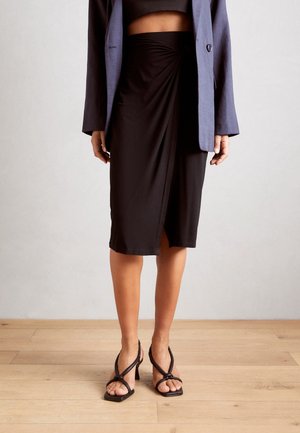 Schwarzer, knielanger Rock mit einem gedrehten Design vorne. Getragen mit einem kurz geschnittenen Oberteil und einem blauen Blazer. Kombiniert mit schwarzen, Riemchen-Absatzschuhen.