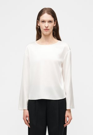 Mujer con cabello castaño liso, vestida con una blusa blanca de manga larga y pantalones negros, de pie contra un fondo blanco liso.