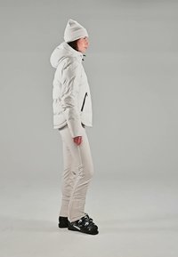Femme en veste d'hiver blanche, pantalon et chapeau, portant des chaussures de ski noires, debout de profil sur un fond gris uni.