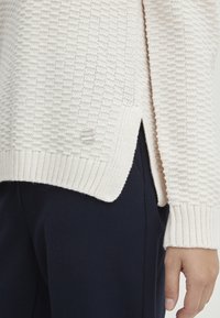 Strickpullover in hellbeige mit strukturiertem Muster, gerippten Bündchen, seitlichen Schlitzen und einem klaren Knopfakzent am Saum.