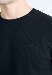 Čierne tričko s textúrou, s okrúhlym výstrihom a jemným embosovaným logom "HUGO" na hrudi. Tkanina má mriežkový vzor, ktorý zvyšuje jeho vizuálny záujem.