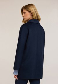 Laivastonsininen villatakki, jossa on lovettu kaulus, pitkät hihat vaaleansinisin hihansuukin ja rento istuvuus. Sileä pinta, jossa on vähäisiä yksityiskohtia.