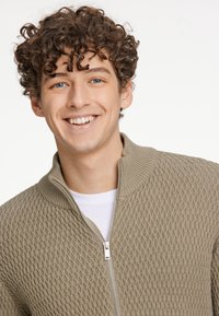 Jersey de punto texturizado beige con cuello alto y cremallera plateada, usado sobre una camisa blanca. Presenta un patrón de rombos y bordes acanalados.