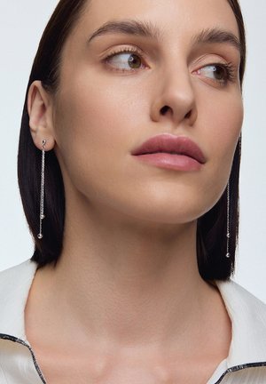 Srebrne kolczyki w kształcie kropli z detalami w postaci łańcuszka i małymi, okrągłymi koralikami. Smukły design na jasnym tle, noszone przez modelkę.