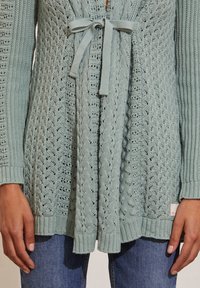 Cárdigan de punto azul claro con patrones texturizados, atado a la cintura con un lazo, usado sobre jeans azules con las manos descansando a los lados.