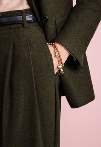 Olive green tailored suit with a navy leather belt, featuring a gold charm bracelet. The texture appears smooth and refined.