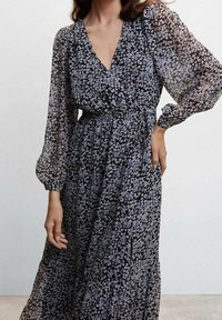 Florale maxi-jurk van doorschijnende zwarte stof met een V-hals, lange pofmouwen en een getailleerde taille. Heeft een gerimpelde zoom en kleine lichtgekleurde bloemen.