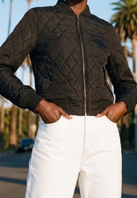 Personne portant une veste matelassée noire zippée et un pantalon blanc, les mains dans les poches, en plein air avec des palmiers en arrière-plan.