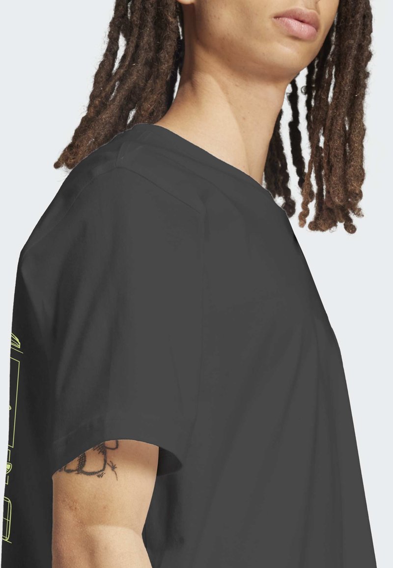 Persoon met dreadlocks, gekleed in een zwart T-shirt met korte mouwen, waarop een gedeeltelijke tattoo op de bovenarm zichtbaar is, tegen een lichte achtergrond.