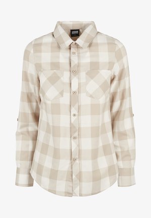 Person iført beige ternet skjorte med opklistrede ærmer, lyseblå bootcut jeans og sorte kraftige sko stående på grå baggrund.