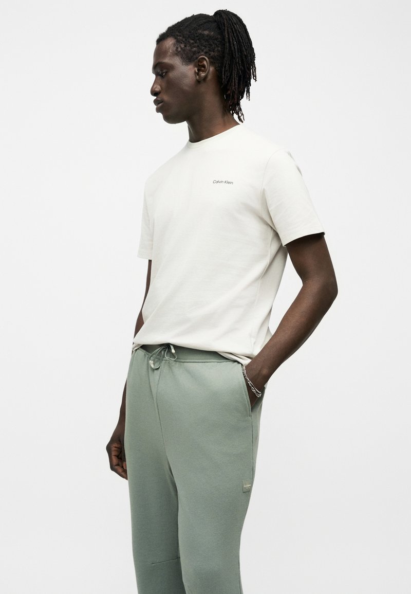 Homme avec des dreadlocks portant un t-shirt blanc Calvin Klein et un pantalon vert clair à cordon, debout avec une main dans la poche.